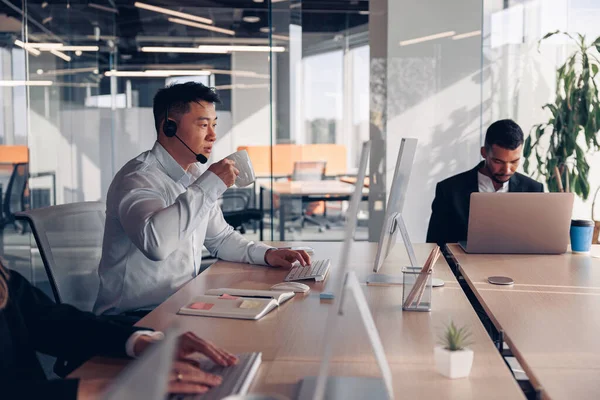 アジアのビジネスマンが同僚を背景にオフィスでコンピュータに取り組んでいる間にコーヒーを飲む — ストック写真