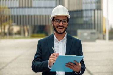 Resmi takım elbiseli gülümseyen Hintli mühendis ve elinde kağıt klasör olan sert şapka. 