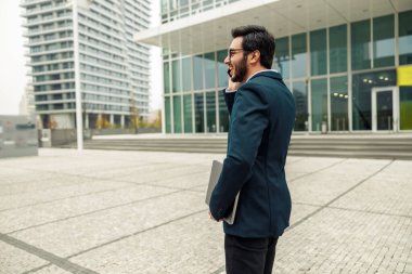 Bilgisayarlı gülümseyen işadamı modern ofis binasının yakınında telefonla konuşuyor. 