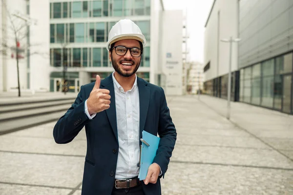 Indian engineer in formal suit and hard hat with paper folder in hands showing thumb up
