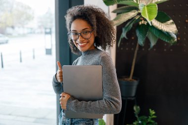 Sırıtkan Afrikalı iş kadını, rahat bir iş yerinde pencerenin yanında dururken dizüstü bilgisayarını tutuyor.
