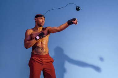 Man with naked torso uses boxing ball simulator for training on studio background with color filter