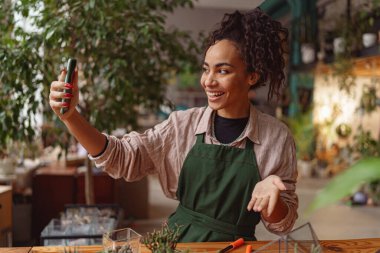 Smiling woman florist taking picture with her plants in own florar studio. Blurred background