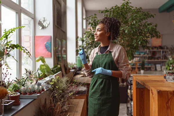 Attractive smiling florist stands on background of her own floral decor studio and looking at window