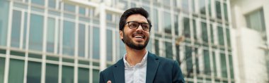 Modern ofis binasının yanında dizüstü bilgisayarlı gülümseyen işadamı yan bakıyor.