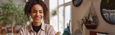 Attractive smiling florist stands on background of her own floral decor studio and looking at camera