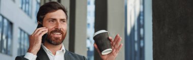 Smiling businessman in suit with coffee talking phone near office building. Business concept