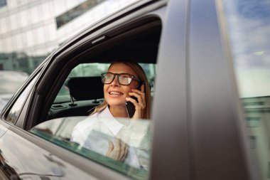 İş kadını arabanın arka koltuğunda seyahat ediyor ve pencereye bakarken akıllı telefonlardan konuşuyor.