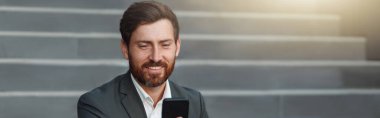 Businessman in suit sitting on stairs with coffee during break and using phone. Blurred background