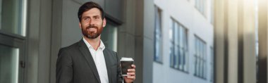 Confident businessman in suit with laptop and coffee near office building. Blurred background