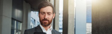 Attractive businessman holding phone standing near office building during break. Business concept