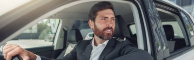 Handsome businessman in grey suit is riding behind steering wheel of car. Blurred background