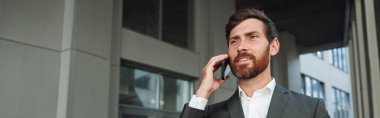 Smiling businessman in suit with coffee talking phone near office building. Business concept