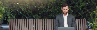 Male businessman working on laptop at lunchtime sitting on bench near office. Blurred background