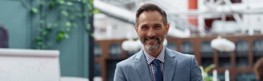 Handsome mature businessman standing in modern office on colleagues background and looking camera