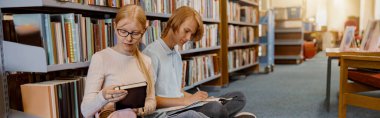 Friends student sit on floor near bookshelves in library and studying. Education concept