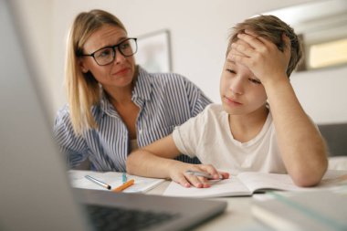 Annesi ona ödevlerinde yardım ederken yorgun, üzgün çocuk dizüstü bilgisayarla masada oturuyor. Yüksek kalite fotoğraf