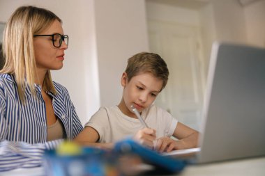 Odaklanmış küçük okul çocuğu annesinin yardımıyla okul ödevlerini yapmak için dizüstü bilgisayar kullanıyor. 