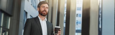 Attractive businessman with coffee and laptop near office building during break. Blurred background