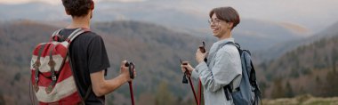 Cheerful woman with walking sticks standing on peak of munt with boyfriend or friend and smiling.