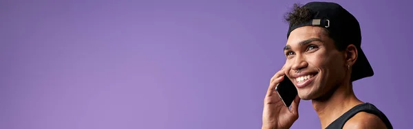 黒のTシャツと帽子でモバイルで話しているトランスジェンダーのブルネットの男の肖像 紫の背景に電話で話すラテン系のトランスジェンダーモデル デジタル技術による人々のライフスタイル — ストック写真