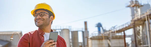 Happy industrial worker, standing with laptop and mobile phone on his hands and looking away at construction industry production plant