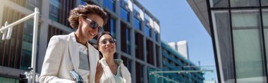 Women in trendy outfits laugh and walk outside near business center. Blurred background