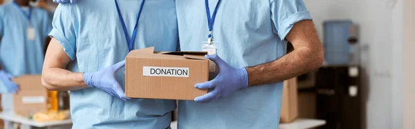 Cropped shot of two caucasian guys, male volunteers in blue uniform, protective gloves posing together and holding cardboard box for Donation. Charitable foundation concept