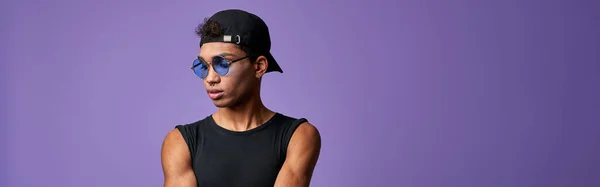 カジュアルな服装でトランスジェンダーの若者の肖像画 ブラックのTシャツとキャップのラテンアメリカのトランスジェンダーモデルは ブルーのジーンズとサングラス 紫の背景に筋肉スポーツマン — ストック写真