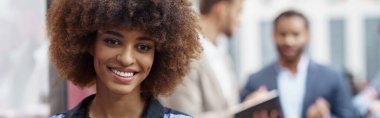 Smiling african businesswoman standing in modern office on colleagues background and looking camera