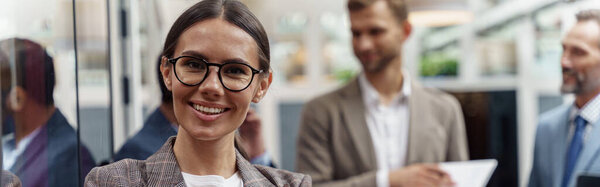 Smiling businesswoman standing in modern office on colleagues background and looking camera