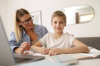 Küçük gülümseyen okul çocuğu annesinin yardımıyla okul ödevlerini yapmak için dizüstü bilgisayar kullanıyor. 