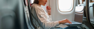 Uçağın camında oturan ve taşınabilir defter üzerinde çalışan genç bir kadın.