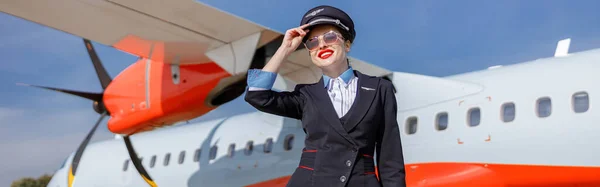 Joyful female flight attendant in airline uniform touching cap and smiling while standing near aircraft at airfield