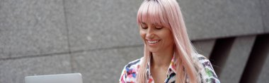 Smiling female with dyed hair clicking smartphone while sitting at table outdoor before laptop