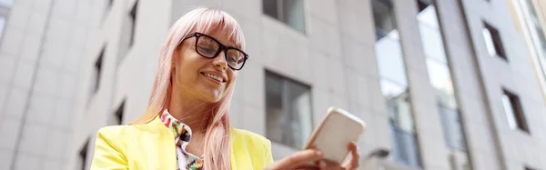 Joyful lady in glasses holding modern smartphone and smiling while standing on the street