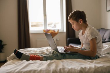 Genç çocuk yatağa uzanıyor ve açık bir dizüstü bilgisayara bakıyor. Evde eğitim kavramı. Yüksek kalite fotoğraf