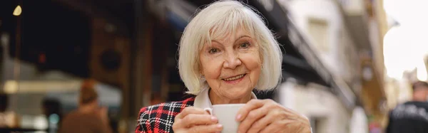 stock image Senior urban lady wearing stylish jacket expressing gladness while tasting mug of beverage in cozy cafeteria