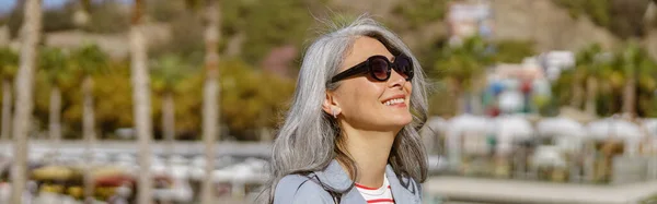 Długowłosy Dojrzała Kobieta Okularach Przeciwsłonecznych Płaszczu Trench Trzymając Smartfona Uśmiechając — Zdjęcie stockowe