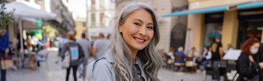 Good looking Asian woman with long grey hair in trench coat holding camera and smiling on street of old town