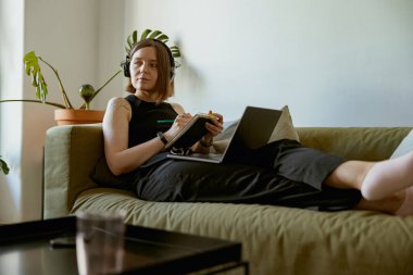 İnternette öğrenmek için dizüstü bilgisayar kullanan ve evdeki kanepede yatarken not alan bir kadın.