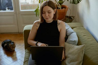 Evdeki rahat koltukta uzanan ve arka planda kedisi olan bir kadın. Mesafe çalışma kavramı 