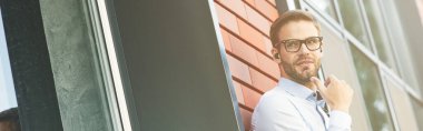 Genç yakışıklı iş adamı, dışarıda beklerken elinde akıllı telefonlar olan kablosuz kulaklıklar takıyor. Modern teknolojileri kullanan iş insanları kavramı