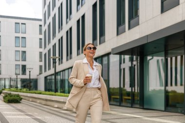 Kahveli gülümseyen kadın ofis çalışanı, teneffüste modern bina arka planında yürüyor.