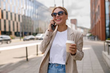 Smiling woman office worker is talking by phone while standing on modern building background 