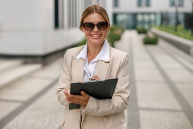 Gülümseyen kadın ofis çalışanı modern binanın arka planında duruyor ve notlar alıyor.