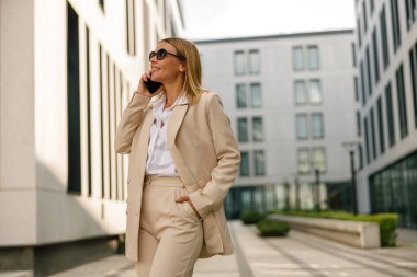 Gülümseyen kadın ofis çalışanı modern binanın arka planında duruyor ve telefonla konuşuyor.