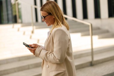 Gülümseyen kadın ofis çalışanı tatilde modern bina arka planında telefon tutuyor.