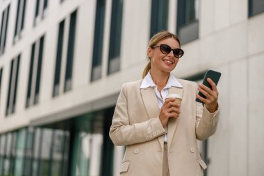 Gülümseyen kadın ofis çalışanı modern binanın arka planında telefon kullanıyor ve kahve içiyor.
