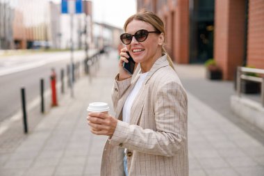 Smiling woman office worker is talking by phone while standing on modern building background 